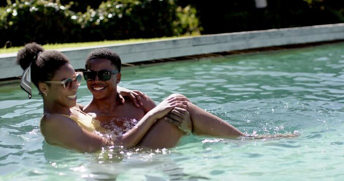 African American couple playing in pool, man holding adjusting woman in yellow swimsuit headscarf