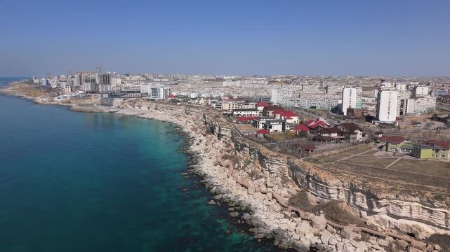 Breathtaking drone ascent revealing a vast panorama of Aktau city stretching to the horizon. The footage features the unique wooden Rock Trail boardwalk winding along the rugged Caspian Sea cliffs. 