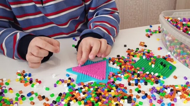 Close-up of a child's hands placing colorful plastic fusebeads onto a pegboard to form a pattern. This activity supports fine motor skill developmentin early childhood.