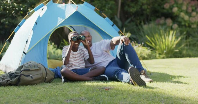 African American father and son at blue tent, father pointing, son scanning with binoculars on lawn