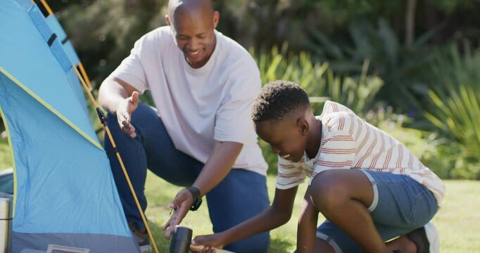 African American father pointing, boy hammering stake with mallet, securing blue tent in yard