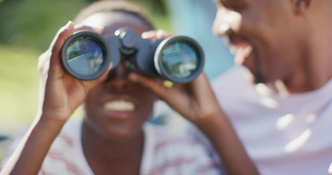 African father and son sharing binoculars, guiding while son peering to spot sunlit house in yard
