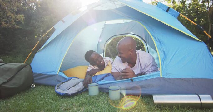 Father and teenage son waking, sunlight streaming into blue tent, laughing and tussling over pillow