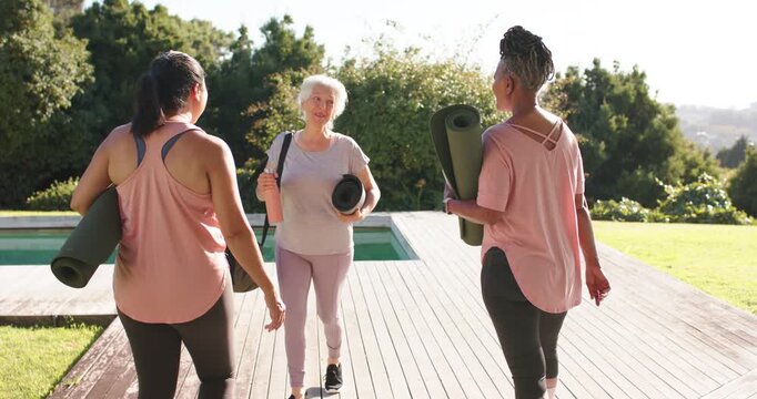 Diverse female friends arriving on deck by pool carrying mats touching face, pointing view for yoga