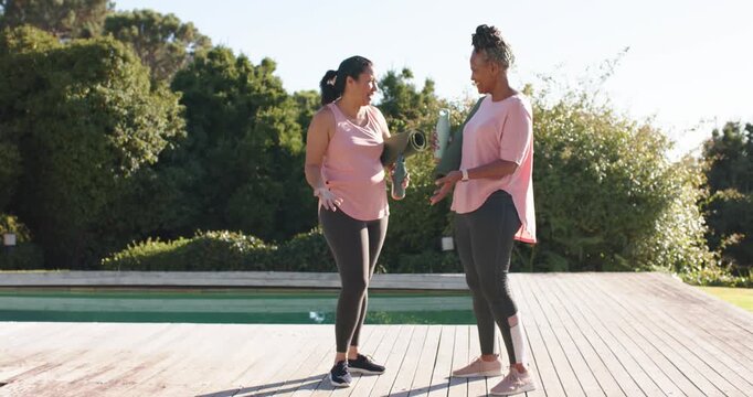 African American women laughing, holding green mats and bottle on deck by pool preparing yoga