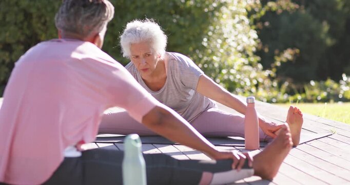 Diverse women stretching, senior leading, mirroring to deepen stretch on deck with mats and bottles