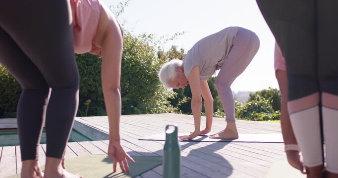 Female group bending forward on sunlit deck near green bottle, senior woman hopping back into plank