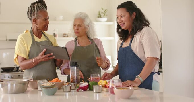 Diverse female friends consulting tablet at kitchen island, agreeing recipe steps arranging bowls