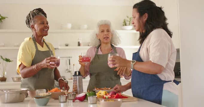 Talking 3 women with pink drinks pointing at veggies on kitchen island, clinking glasses in aprons