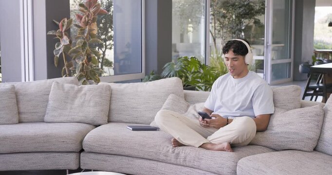 Adult man scrolling phone, reaching for notebook and writing notes on sofa while wearing headphones