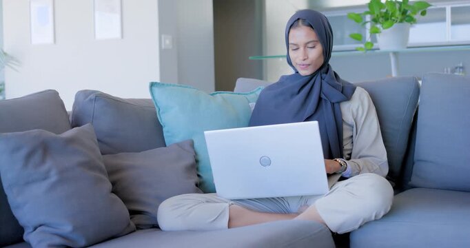 Woman in dark hijab responding to laptop, typing, pausing and glancing while studying on gray sofa
