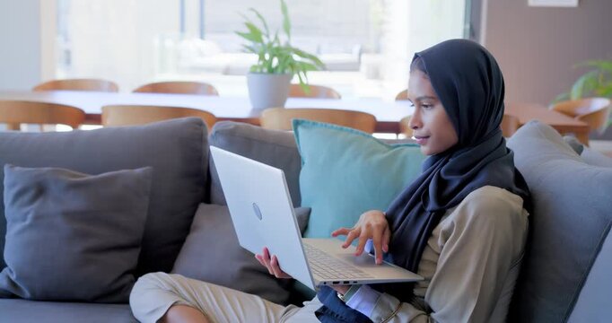 Woman in dark hijab pausing then typing and scrolling on silver laptop at home to work