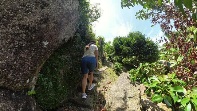 Woman Climbing Steep Rocky Steps Surrounded by Vertical Jungle Walls and Bright Sunlight Breaking Through, Low Angle View, Tropical Trekking Concept