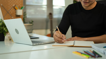 Focused business man working on laptop computer making notes sitting at home office desk. Young adult male professional manager doing marketing research writing checklist on paper at workplace
