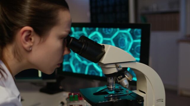 POV handheld shot of female Caucasian researcher in white coat analyzing cells via microscope and AI computer software while male Black colleague working in background in smart laboratory