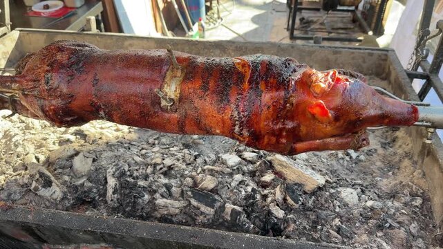 Whole suckling pig slowly roasting on a rotating spit over hot charcoal. The skin is golden-brown and crispy, capturing a traditional outdoor rustic culinary process