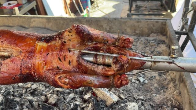 Whole suckling pig turns on a rotisserie spit over glowing coals. The crackling golden skin and rising heat showcase the authentic art of slow-roasting meat at a traditional outdoor feast