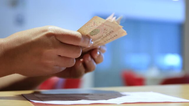 Hands counting Thai banknotes while pointing at a document on a desk, representing financial planning, payment, budgeting, money, cash, and personal expense management