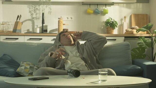 Close-up of a man in a hoodie sitting at home and reaching for pills on the table. Thoughtful or tired expression, alarm clock in the foreground. Soft daylight, domestic interior. Concept of stress