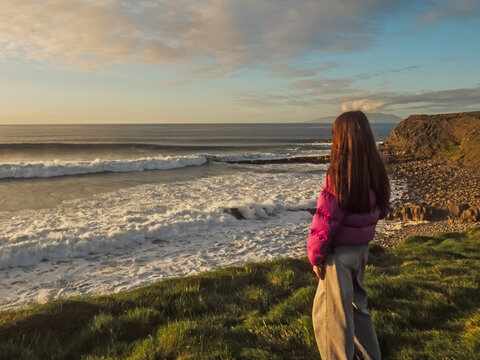 Woman stands on grassy cliff near ocean waves. Red jacket contrasts with blue sky and sea. She smiles gently, facing horizon with calm. Sunlight highlights her hair and silhouette