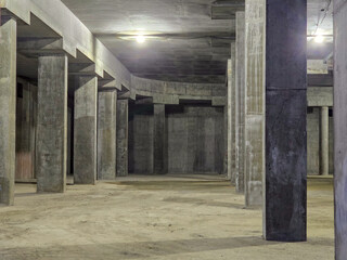 Concrete pillars stand tall under dim lighting. Floor shows signs of construction and neglect. Space feels empty, echoing with industrial strength. Image evokes raw, unfinished potential © Антон Скрипачев