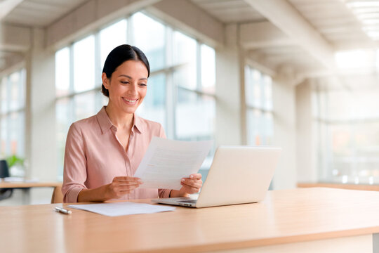 Businesswoman reviewing a financial report and working on her laptop inside a modern office. Entrepreneur analyzing documents with focus and a professional attitude, dedicated to her work goals