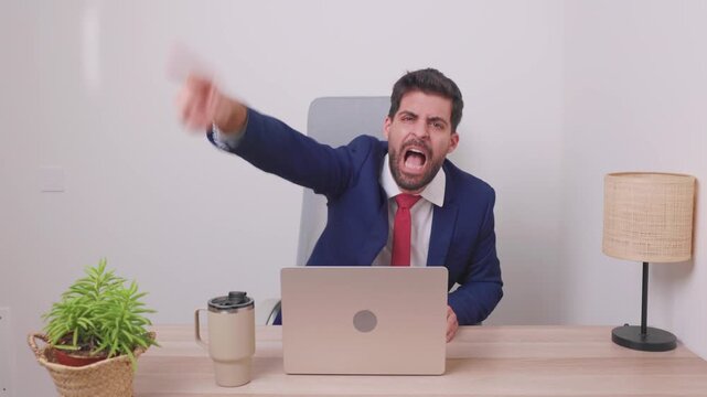 Frustrated boss in suit shouting and pointing at laptop camera during tense video conference, angrily gesturing dismissal while working remotely from his office, accusing employee
