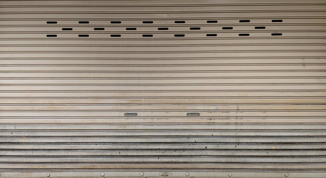 Gray metal roller shutter door. Industrial gray roller shutter door with ventilation slots. Authentic weathered metal shutter door with subtle usage marks. Warehouse or commercial building background.