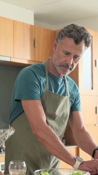 Vertical video: Pouring glass offered, man in apron preparing salad at counter with bowl and knife