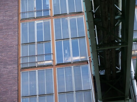 Medium shot of a weathered industrial facade featuring large multi-pane
windows with orange and green metal frames. A green industrial skybridge with
thick pipes runs vertically through 