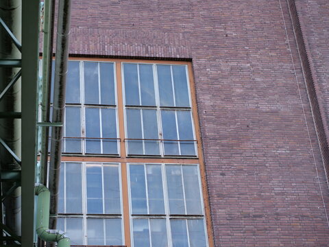 Medium shot of a weathered industrial facade featuring large multi-pane
windows with orange and green metal frames. A green industrial skybridge with
thick pipes runs vertically through 