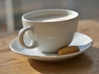 Obraz premium Close-up, side view of a white ceramic cup filled with frothy cappuccino or latte, placed on a matching saucer. A small, golden-brown cookie sits on the side of the saucer. The set rests on a light