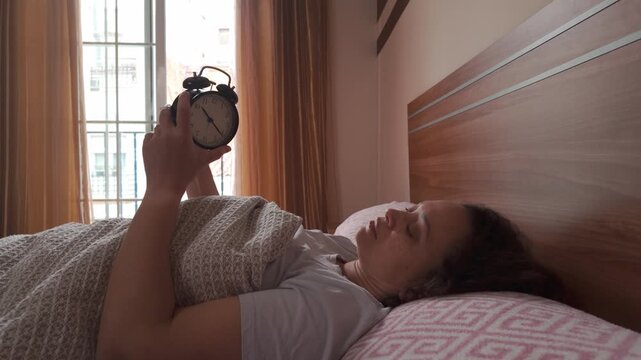 Young woman adjusts her alarm clock while waked up in the morning, still lying on the bed