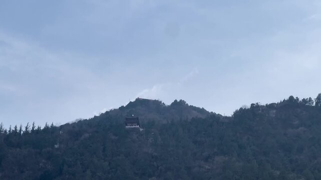 Sunlit winter landscape of a historic mountain hot spring site in China, featuring traditional Chinese architecture nestled among forested hills. Visitors gather around a serene pavilion and water fea