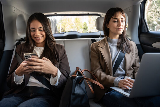 Businesswomen working in car backseat with phone and laptop