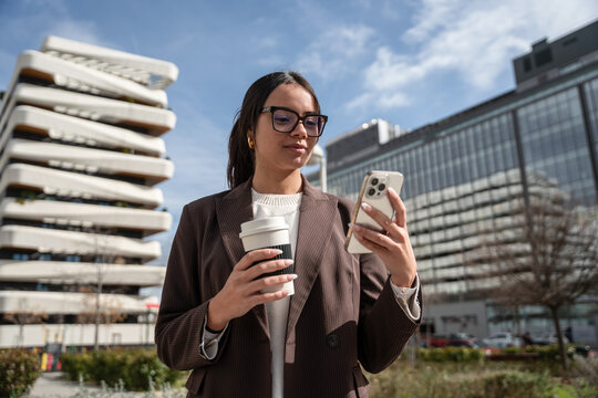 Businesswoman checking smartphone while holding coffee in city
