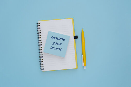 A spiral notebook with a blue cover and a yellow pen on a light blue background. A sticky note reads 'Assume good intent'.