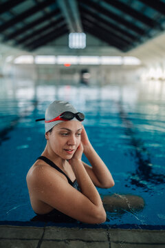 Young woman swimmer with goggles and swim cap by the pool edge in an indoor stadium, preparing for training
