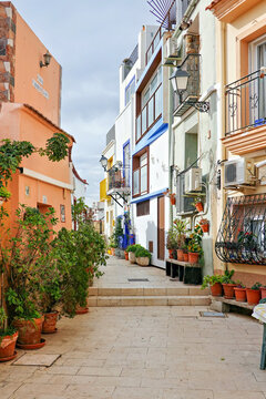 Narrow cozy street with white houses, flowers in potted plants in historic Santa Cruz neighborhood of Alicante Old Town, located near Santa Barbara Castle