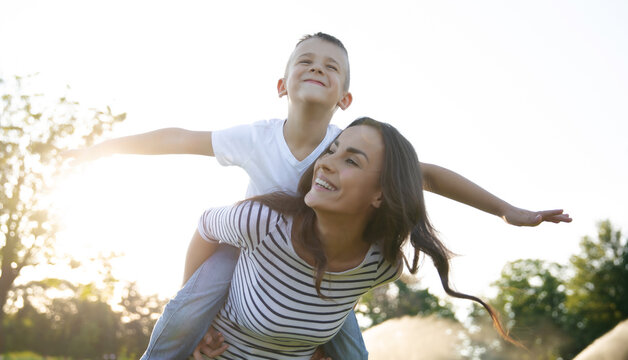 Smiling mother giving her son a piggyback ride outdoors, playful family moment, child pretending to fly, summer fun and Mother&rsquo;s Day concept.
