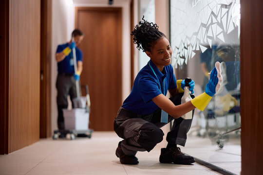 Black janitorial service employee polishing glass surface in office building.