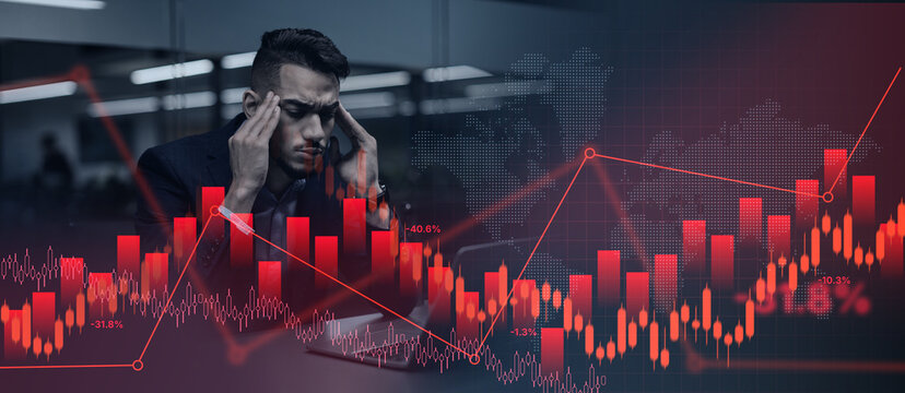 A businessman sits at a desk in an office. He holds his head in frustration while looking at charts and graphs. The background shows stock market data with red trends.