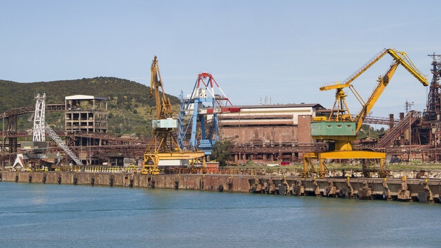 View of industrial cranes and aged ironworks stand in stark contrast to the calm turquoise sea under a clear sky, Piombino, Tuscany, Italy.