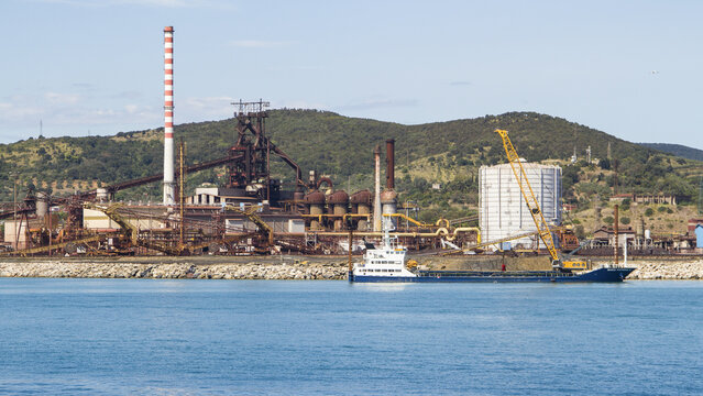 View of the industrial complex with its towering chimney and cranes contrasts with the calm blue sea and distant green hills, Piombino, Tuscany, Italy.