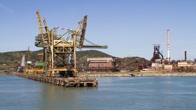 View of a large industrial port with cranes reflected in the still turquoise water, backed by hills and a clear sky, Piombino, Tuscany, Italy.