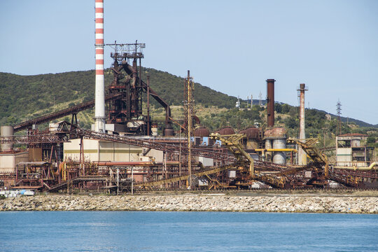 View of a sprawling industrial complex with towering chimneys and rusted metal structures meets the serene blue sea, contrasting industry with nature, Piombino, Tuscany, Italy.