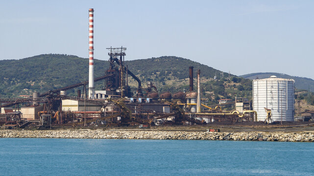 View of the industrial complex with a towering chimney, juxtaposed against the serene sea and distant hills, creating a striking contrast, Piombino, Tuscany, Italy.