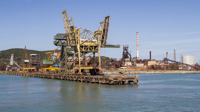View of a colossal, weathered crane looming over the tranquil, cerulean waters, juxtaposed against the industrial skyline, Piombino, Tuscany, Italy.
