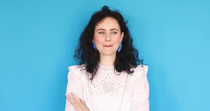 Video of happy coy young woman in white lace blouse hugging herself and then playfully shrugging with smile isolated on blue background, expressive female portrait of joy and flirtation