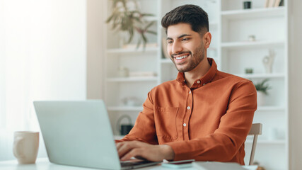Fototapeta premium A young man smiles while typing on a laptop at a table. He is in a bright room with plants in the background. A coffee cup is on the table beside him.